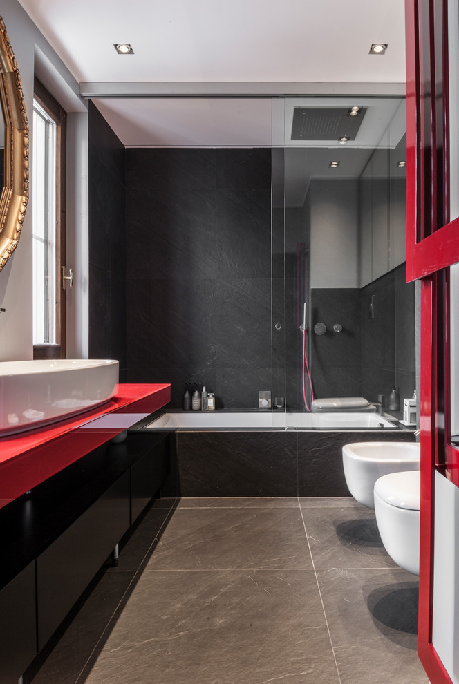 Cette image montre une salle de bain principale design avec un sol en carrelage de céramique, une vasque, un plan de toilette en verre, un placard à porte plane, des portes de placard noires, une baignoire encastrée, un combiné douche/baignoire, un bidet, un carrelage noir, un sol gris, une cabine de douche à porte coulissante et un plan de toilette rouge.