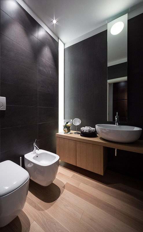 Modernes Badezimmer mit flächenbündigen Schrankfronten, hellen Holzschränken, Wandtoilette, schwarzen Fliesen, braunem Holzboden, Aufsatzwaschbecken und Waschtisch aus Holz in Mailand