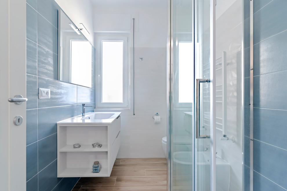 Modern inredning av ett mellanstort vit vitt badrum med dusch, med släta luckor, vita skåp, en hörndusch, en toalettstol med separat cisternkåpa, blå kakel, keramikplattor, vita väggar, ett integrerad handfat och dusch med skjutdörr