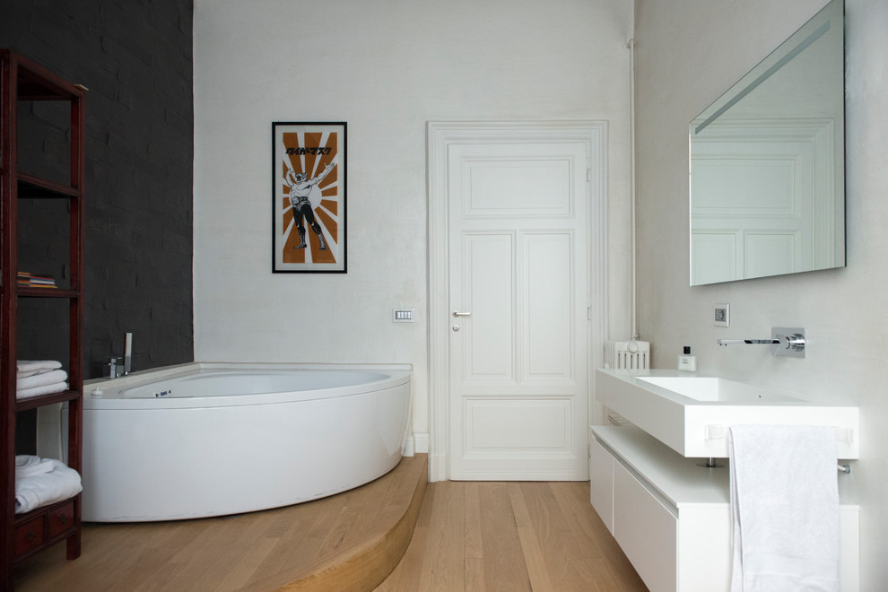 Inspiration för ett funkis vit vitt badrum, med släta luckor, vita skåp, ett hörnbadkar, vita väggar, ljust trägolv, ett väggmonterat handfat och brunt golv