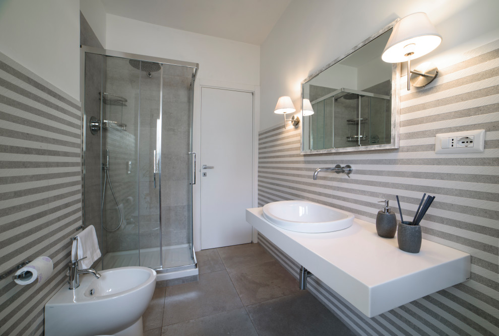 Стильный дизайн: ванная комната среднего размера в современном стиле с белыми фасадами, угловым душем, раздельным унитазом, серой плиткой, керамогранитной плиткой, белыми стенами, полом из керамогранита, душевой кабиной, настольной раковиной, столешницей из искусственного камня, серым полом, душем с раздвижными дверями, белой столешницей, тумбой под одну раковину, подвесной тумбой и панелями на части стены - последний тренд