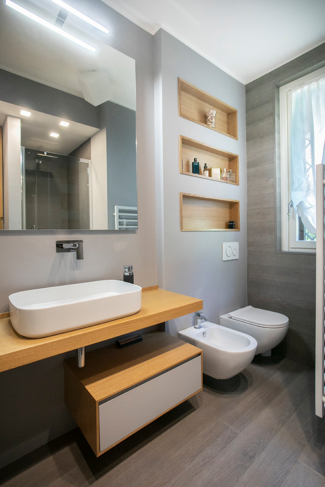 Foto på ett mellanstort funkis beige badrum med dusch, med grått golv, öppna hyllor, grå skåp, en öppen dusch, en toalettstol med separat cisternkåpa, grå kakel, grå väggar, ljust trägolv, ett fristående handfat, träbänkskiva och dusch med gångjärnsdörr