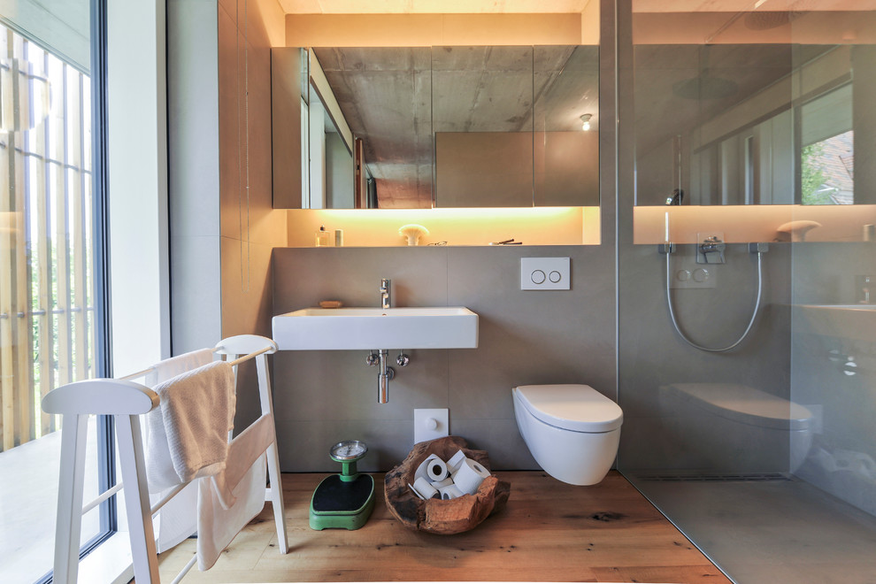 Ispirazione per una stanza da bagno con doccia design di medie dimensioni con doccia a filo pavimento, WC sospeso, piastrelle grigie, pavimento in legno massello medio, lavabo sospeso e pareti grigie