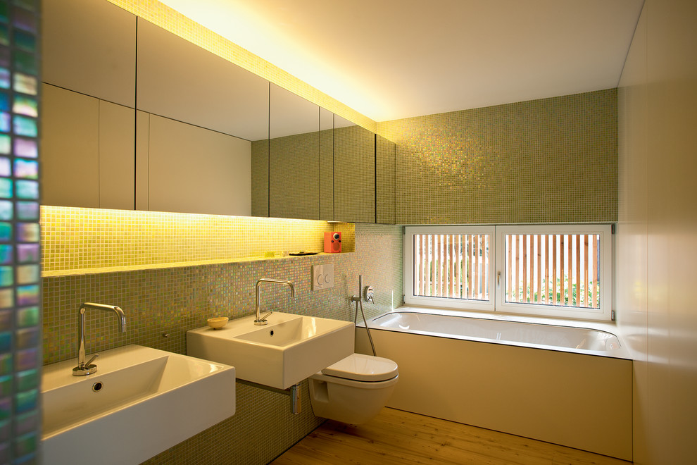 Diseño de cuarto de baño actual pequeño con sanitario de pared, baldosas y/o azulejos verdes, bañera encastrada sin remate, baldosas y/o azulejos en mosaico, suelo de madera clara, lavabo suspendido y paredes verdes