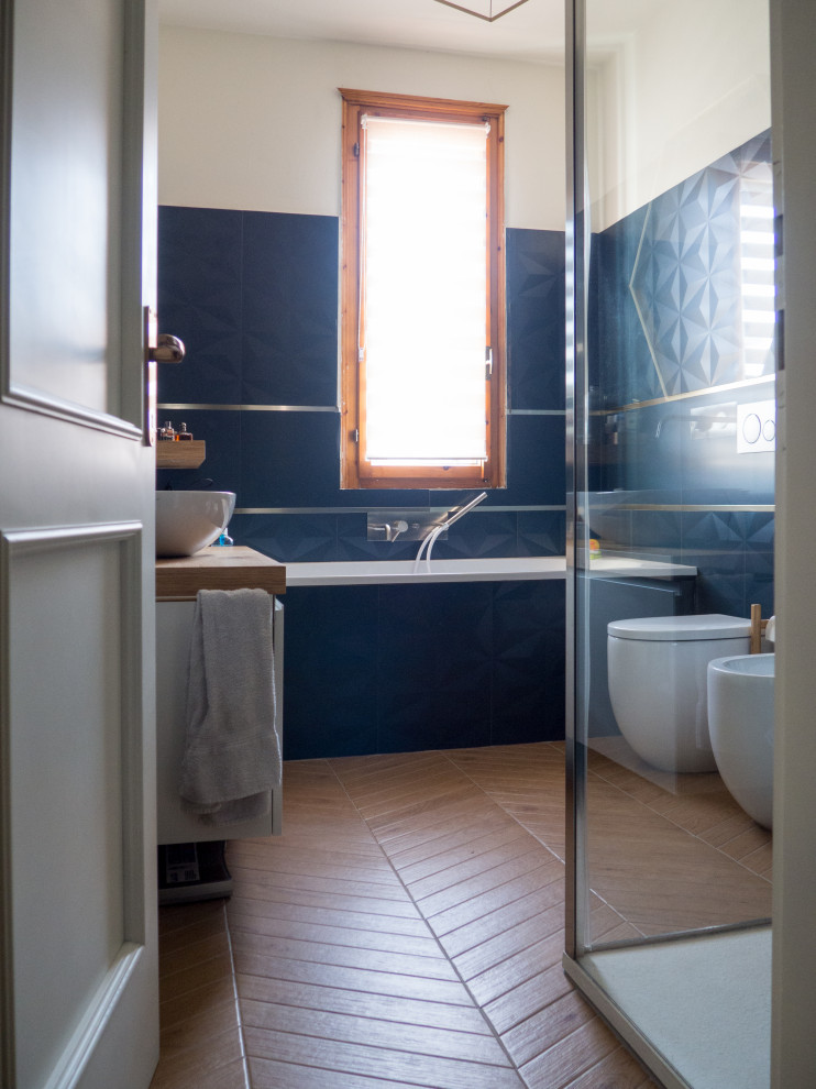 Cette photo montre une grande salle de bain principale tendance avec un placard à porte plane, des portes de placard blanches, une baignoire posée, une douche d'angle, WC séparés, un carrelage bleu, des carreaux de porcelaine, un sol en carrelage de porcelaine, une vasque, un plan de toilette en bois, un sol marron et une cabine de douche à porte coulissante.