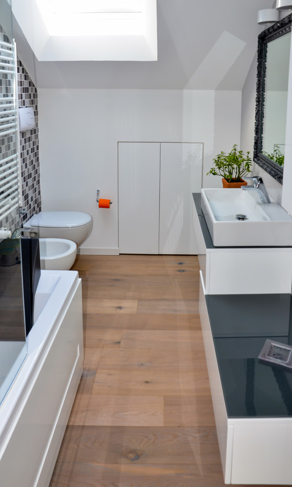 Exemple d'une salle de bain principale tendance en bois clair de taille moyenne avec un placard à porte plane, WC suspendus, un carrelage gris, des carreaux de porcelaine, un mur gris, un sol en bois brun, une vasque et un plan de toilette en verre.