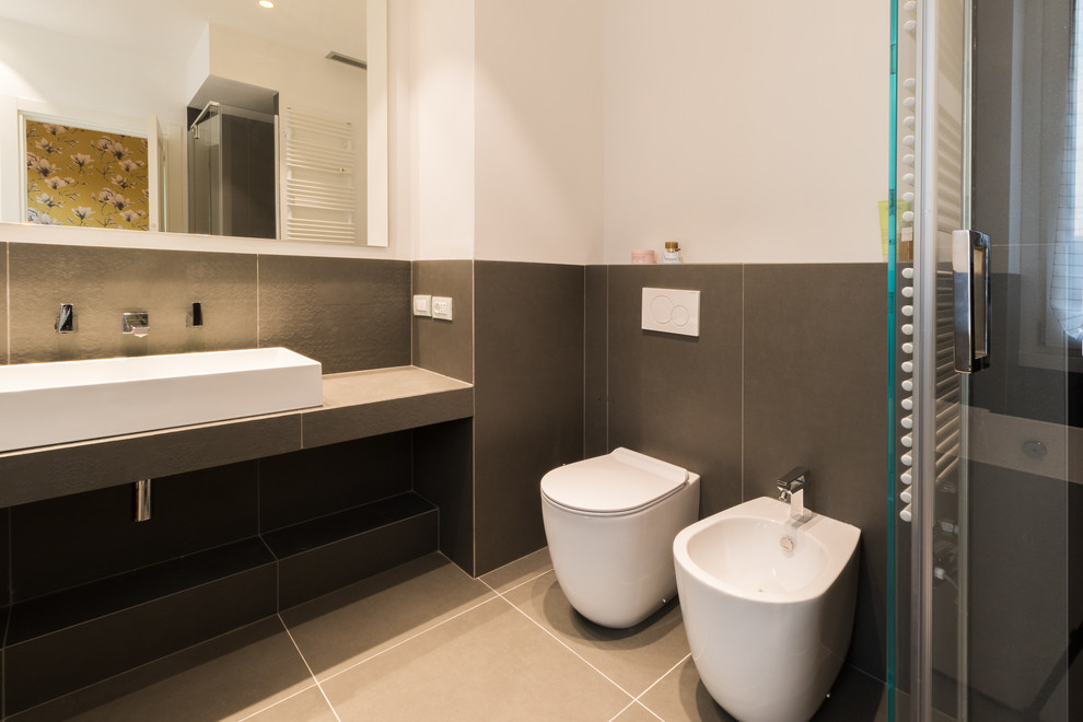Photo of a medium sized contemporary ensuite bathroom in Milan with a freestanding bath, a built-in shower, grey tiles, porcelain tiles, white walls, porcelain flooring, a trough sink, tiled worktops, grey floors, a two-piece toilet and a hinged door.