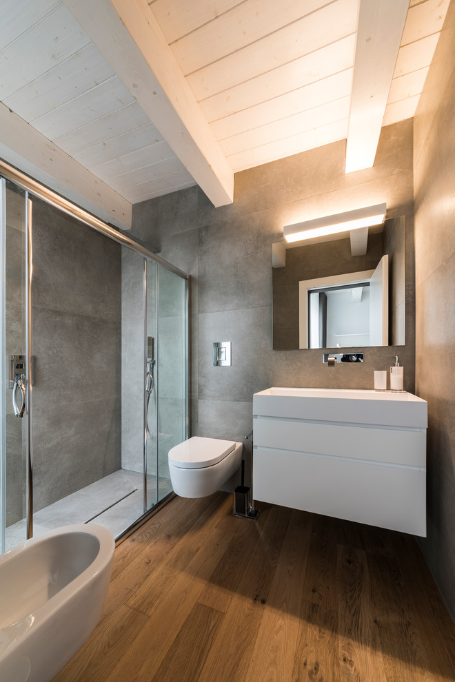 Idéer för att renovera ett mellanstort funkis en-suite badrum, med släta luckor, vita skåp, en dusch i en alkov, en bidé, grå väggar, mellanmörkt trägolv, ett väggmonterat handfat, brunt golv, dusch med skjutdörr och grå kakel