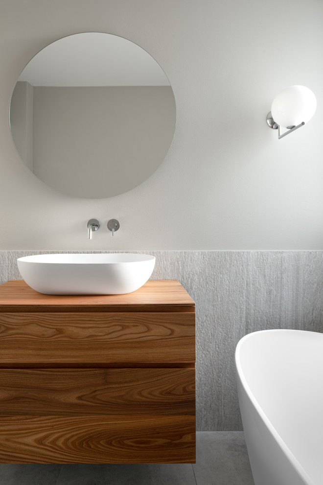 Inspiration pour une salle d'eau design en bois foncé avec un placard à porte plane, une baignoire indépendante, un carrelage gris, un mur gris, une vasque, un plan de toilette en bois et un plan de toilette marron.