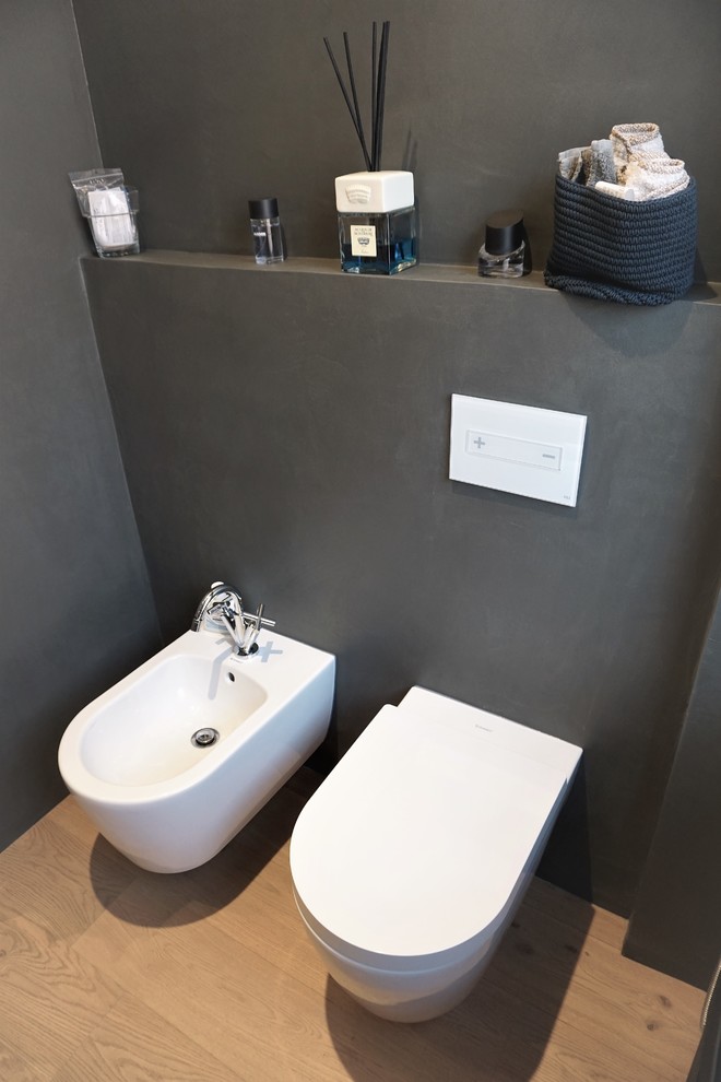 Bild på ett mellanstort funkis toalett, med luckor med profilerade fronter, vita skåp, en vägghängd toalettstol, grå kakel, grå väggar, mellanmörkt trägolv och ett piedestal handfat