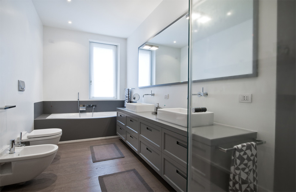 Modern inredning av ett badrum, med grå skåp, ett platsbyggt badkar, en vägghängd toalettstol, vita väggar, mörkt trägolv, ett avlångt handfat och luckor med infälld panel