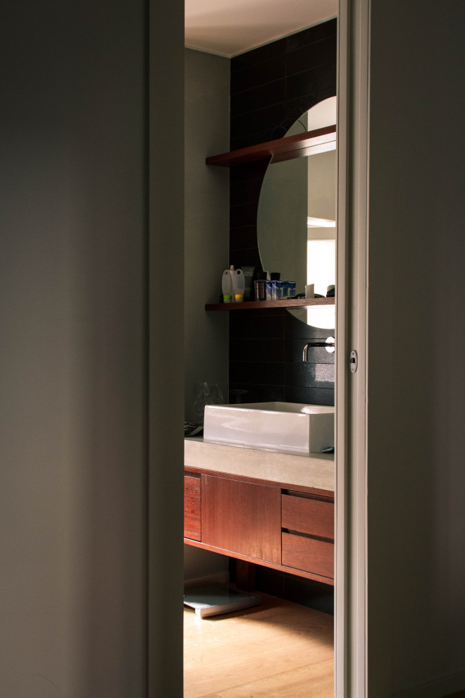 Inredning av ett minimalistiskt mellanstort grå grått badrum med dusch, med möbel-liknande, svarta skåp, en dusch i en alkov, en toalettstol med hel cisternkåpa, svart kakel, marmorkakel, grå väggar, mellanmörkt trägolv, ett fristående handfat, bänkskiva i betong och dusch med gångjärnsdörr