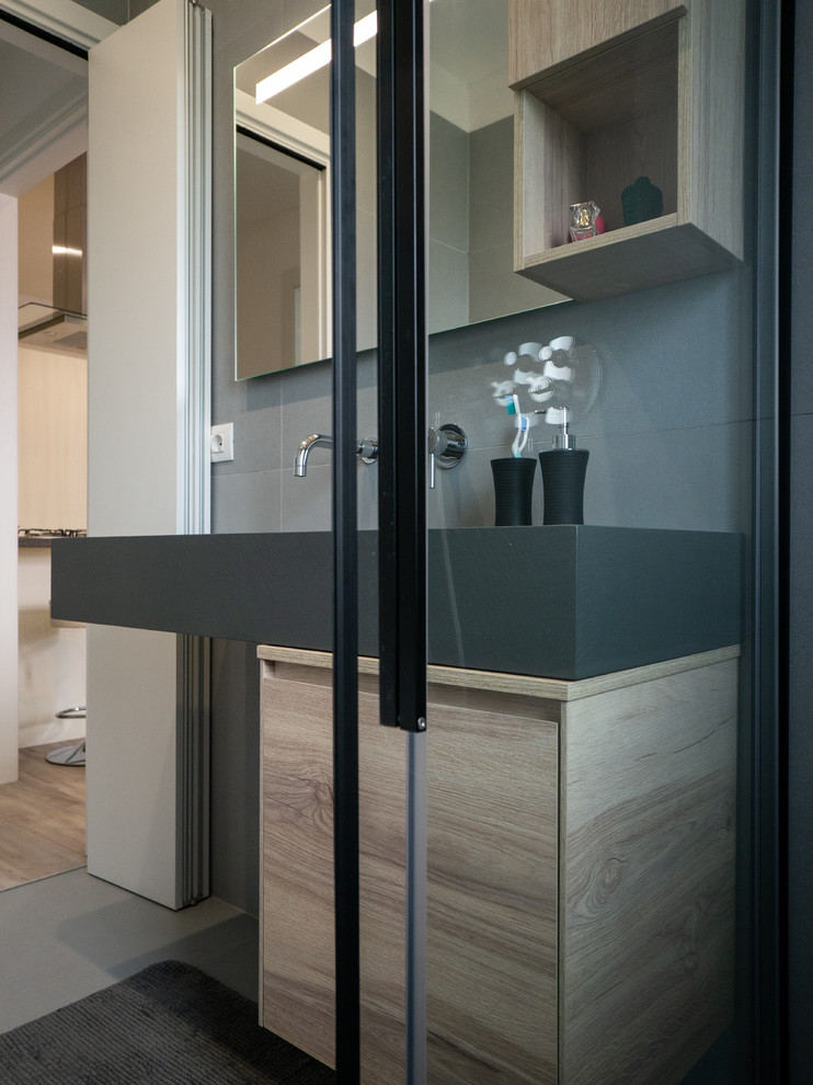 Small contemporary shower room bathroom in Milan with flat-panel cabinets, light wood cabinets, a built-in shower, a wall mounted toilet, grey tiles, porcelain tiles, grey walls, porcelain flooring, a console sink, solid surface worktops, grey floors, a sliding door and grey worktops.