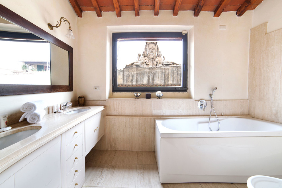 На фото: ванная комната в средиземноморском стиле с плоскими фасадами, белыми фасадами, отдельно стоящей ванной, душем над ванной, бежевой плиткой, белыми стенами, врезной раковиной, бежевым полом, открытым душем и бежевой столешницей