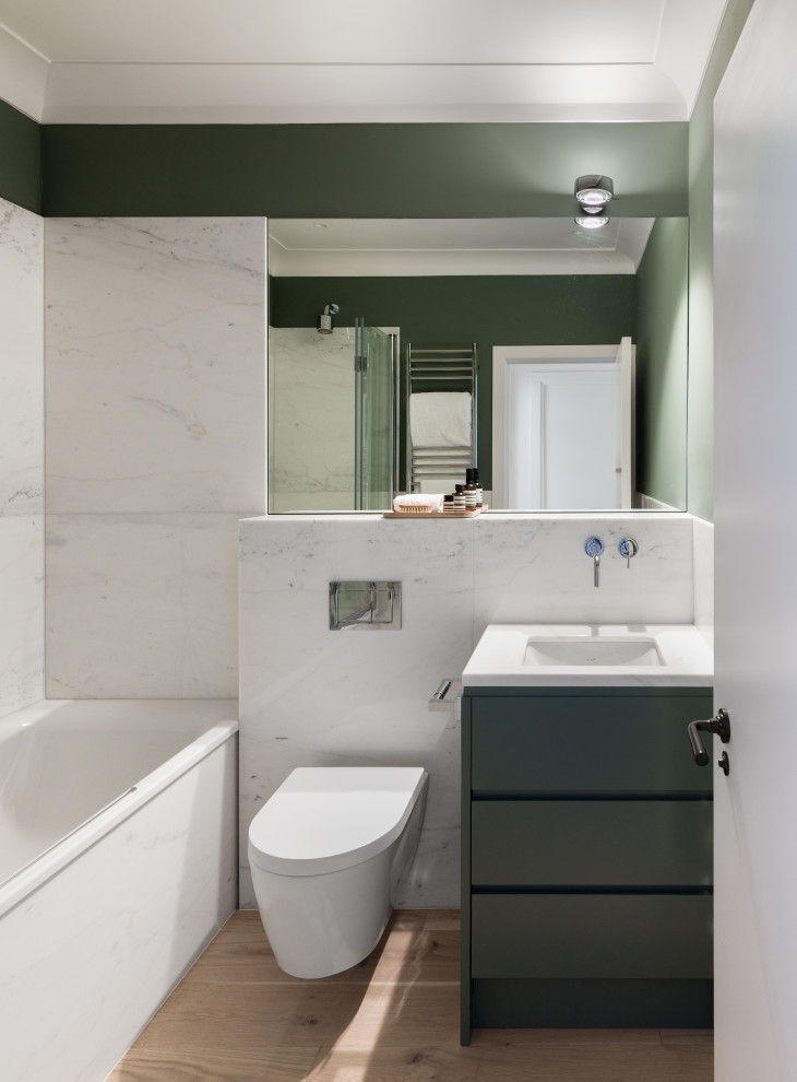 Contemporary shower room bathroom in London with flat-panel cabinets, green cabinets, a built-in bath, a shower/bath combination, a wall mounted toilet, white tiles, green walls, light hardwood flooring, a submerged sink, beige floors and white worktops.