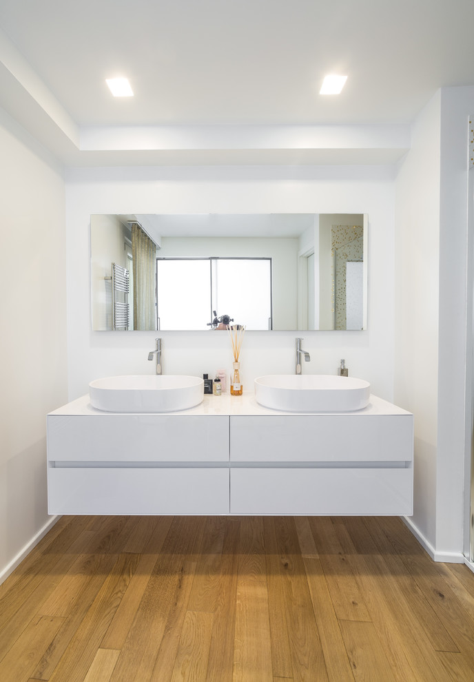 Réalisation d'une salle de bain design avec une vasque, un placard à porte plane, des portes de placard blanches, un mur blanc et un sol en bois brun.