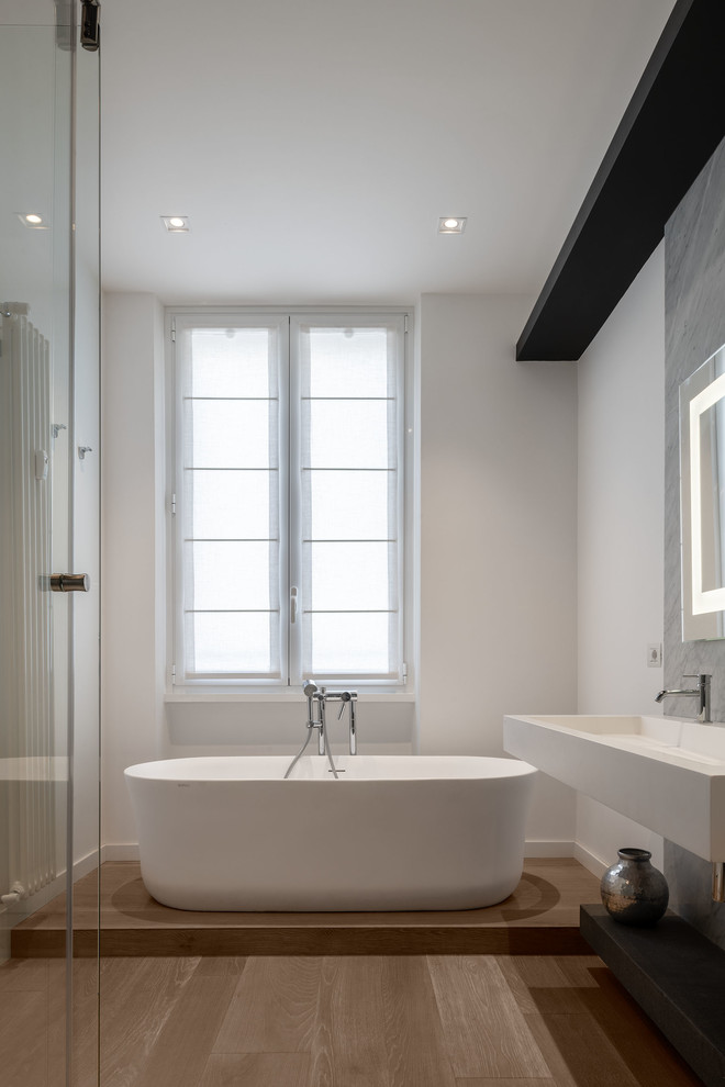 Bild på ett mellanstort funkis vit vitt en-suite badrum, med öppna hyllor, ett fristående badkar, grå kakel, stenhäll, vita väggar, ett väggmonterat handfat, bänkskiva i akrylsten och brunt golv