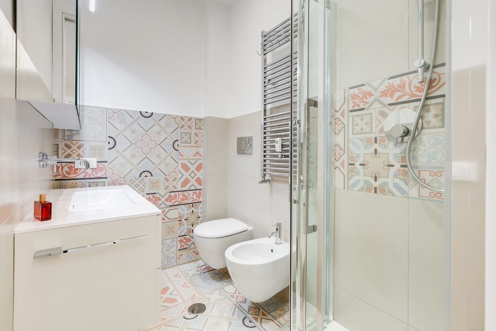 Aménagement d'une salle de bain contemporaine avec un placard à porte plane, des portes de placard blanches, une douche d'angle, un bidet, un mur blanc, un lavabo posé, un sol multicolore et un plan de toilette blanc.
