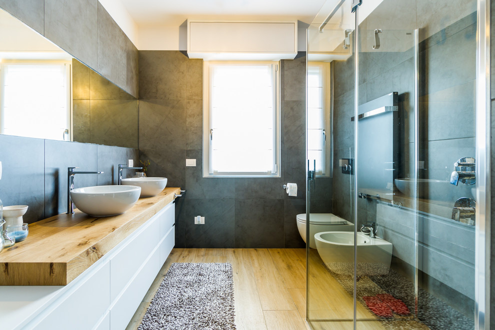 Mid-sized trendy 3/4 gray tile medium tone wood floor and beige floor corner shower photo in Milan with flat-panel cabinets, white cabinets, gray walls, a vessel sink, wood countertops, a wall-mount toilet and a hinged shower door