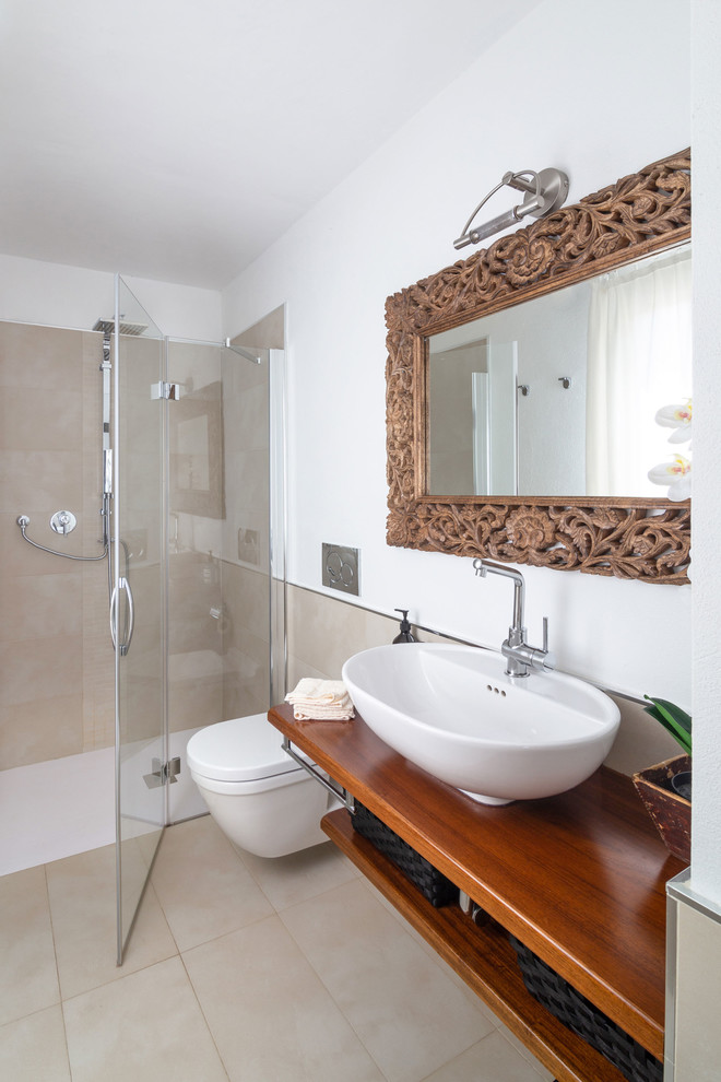 Bild på ett medelhavsstil brun brunt badrum, med öppna hyllor, skåp i mellenmörkt trä, en kantlös dusch, en vägghängd toalettstol, beige kakel, vita väggar, ett fristående handfat, träbänkskiva, beiget golv och dusch med gångjärnsdörr