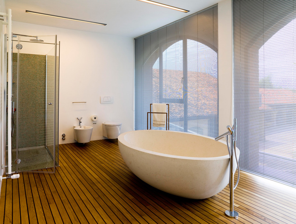Inredning av ett modernt stort badrum, med ett fristående badkar, en hörndusch, en vägghängd toalettstol, vita väggar och mellanmörkt trägolv