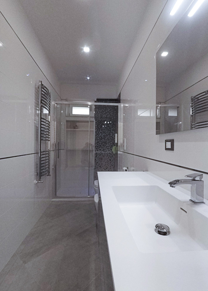 Design ideas for a medium sized contemporary shower room bathroom in Bari with flat-panel cabinets, grey cabinets, a built-in shower, a two-piece toilet, white tiles, porcelain tiles, white walls, porcelain flooring, an integrated sink, solid surface worktops, grey floors, a sliding door and white worktops.