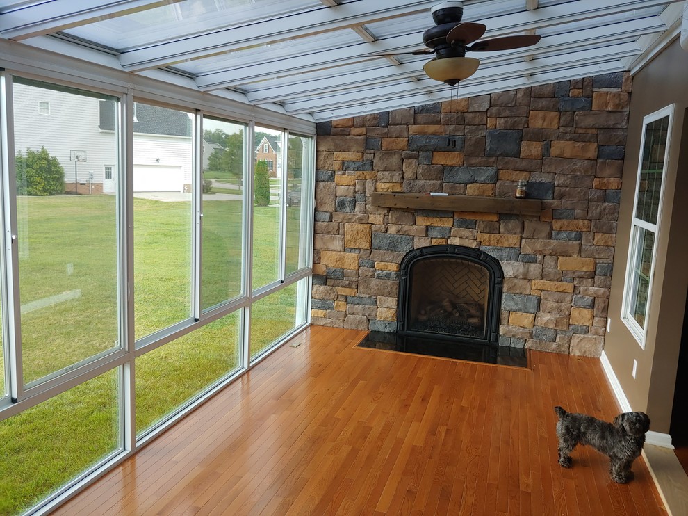 Exemple d'une grande véranda avec parquet clair, une cheminée standard, un manteau de cheminée en pierre, un plafond en verre et un sol marron.