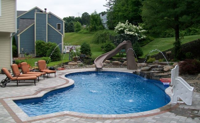 Modelo de piscina con tobogán alargada tradicional de tamaño medio a medida en patio trasero con adoquines de ladrillo