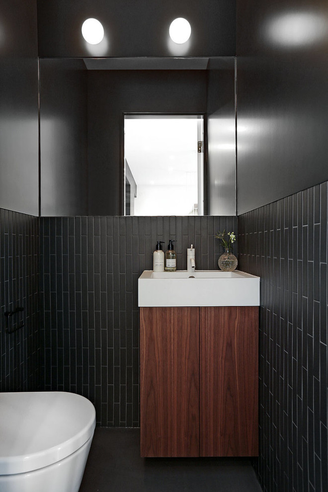 Cette image montre un WC et toilettes minimaliste en bois foncé de taille moyenne avec un placard à porte plane, un mur noir, une vasque, des carreaux de céramique et un sol noir.