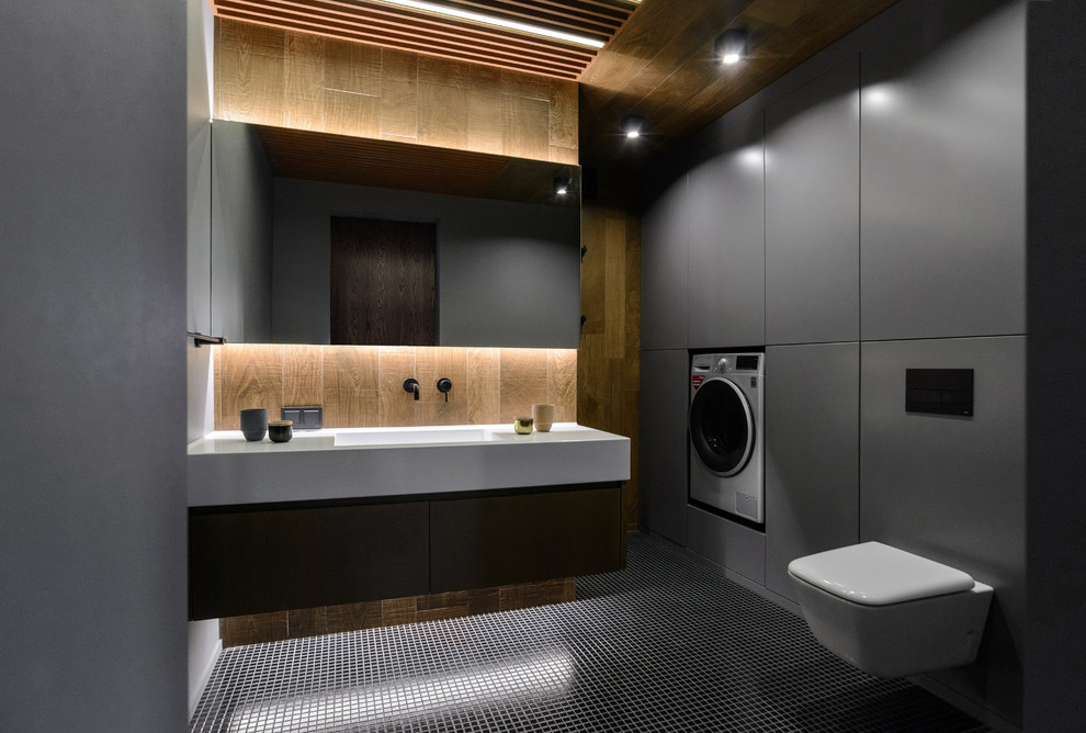 Modelo de aseo contemporáneo con puertas de armario grises, sanitario de pared, lavabo integrado, suelo negro y encimeras blancas