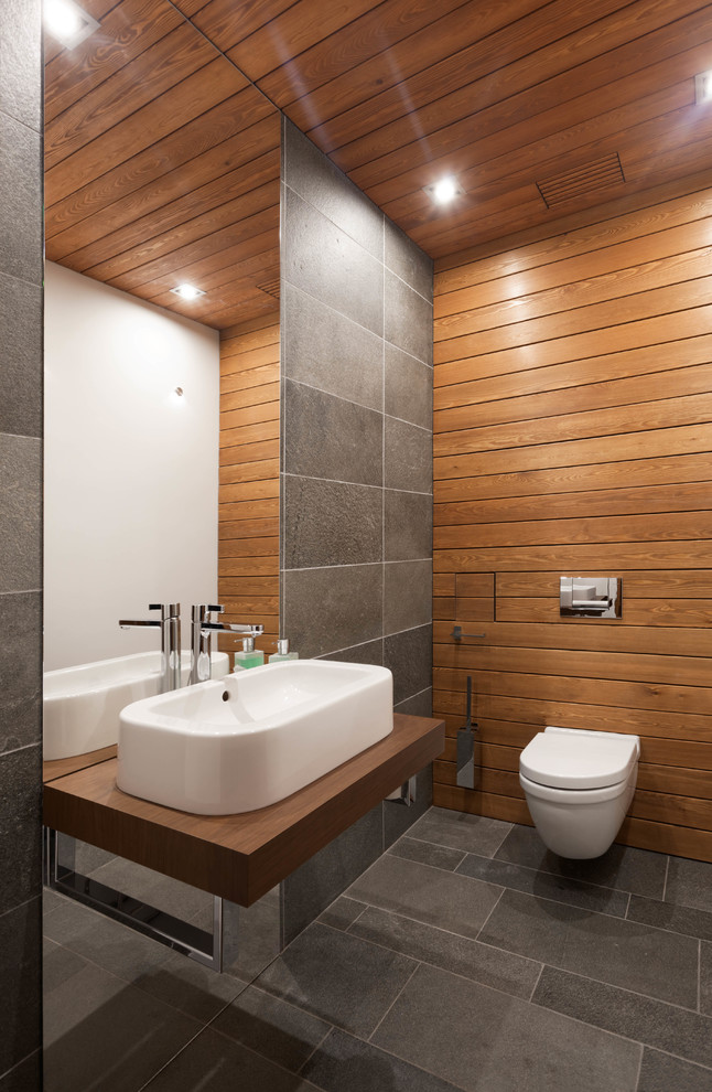 Bild på ett litet funkis brun brunt toalett, med en vägghängd toalettstol, grå kakel, skifferkakel, flerfärgade väggar, skiffergolv, ett fristående handfat, träbänkskiva och grått golv