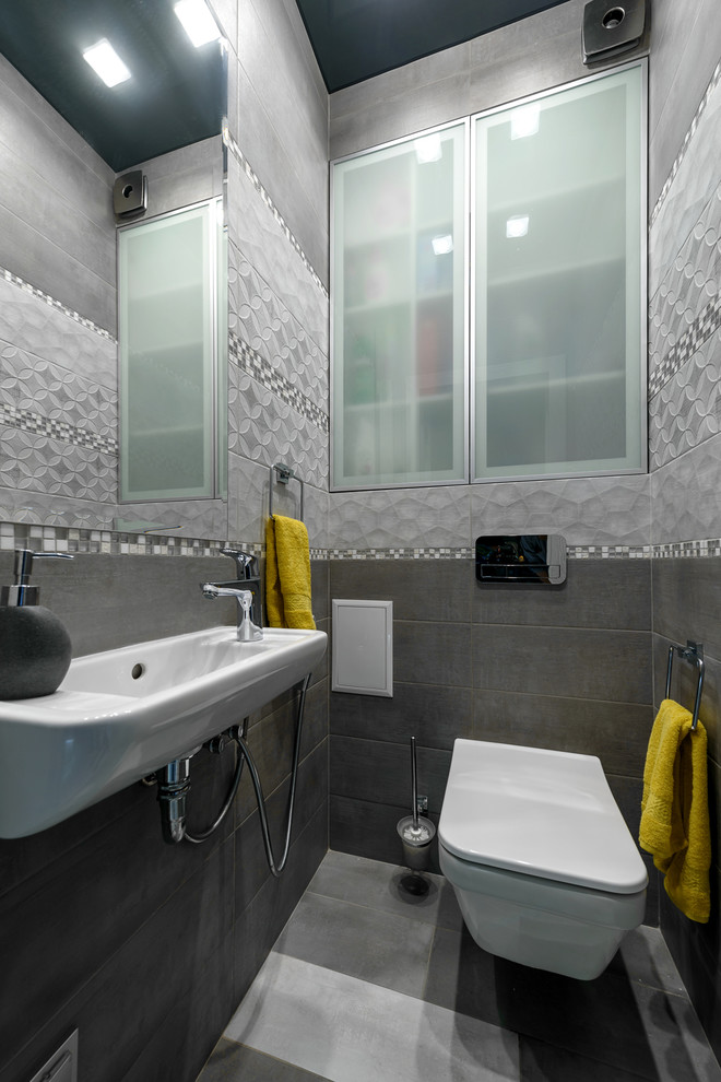 Photo of a small contemporary cloakroom in Other with glass-front cabinets, a wall mounted toilet, grey tiles, porcelain tiles, grey walls, porcelain flooring, a wall-mounted sink and grey floors.