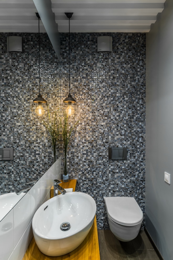 Contemporary cloakroom in Saint Petersburg with a wall mounted toilet, grey tiles, grey walls, a vessel sink, wooden worktops, brown floors and brown worktops.