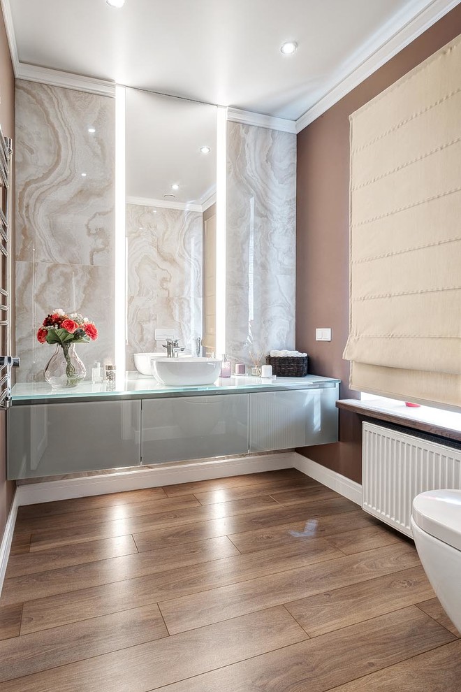 Photo of a medium sized contemporary cloakroom in Moscow with glass-front cabinets, grey cabinets, a wall mounted toilet, beige tiles, porcelain tiles, brown walls, laminate floors, a vessel sink, glass worktops, brown floors and grey worktops.