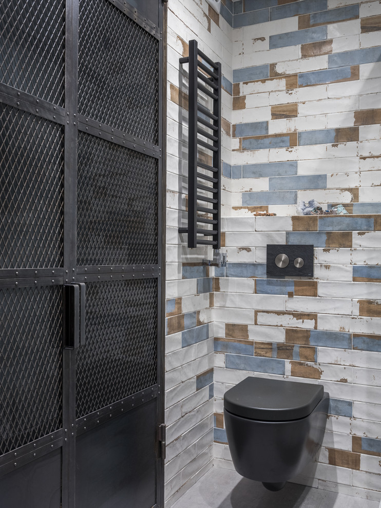 Industriell inredning av ett toalett, med en vägghängd toalettstol, flerfärgad kakel och grått golv