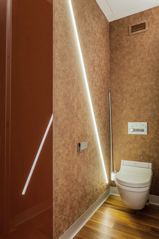 Industriell inredning av ett litet toalett, med en vägghängd toalettstol, bruna väggar, mörkt trägolv och brunt golv