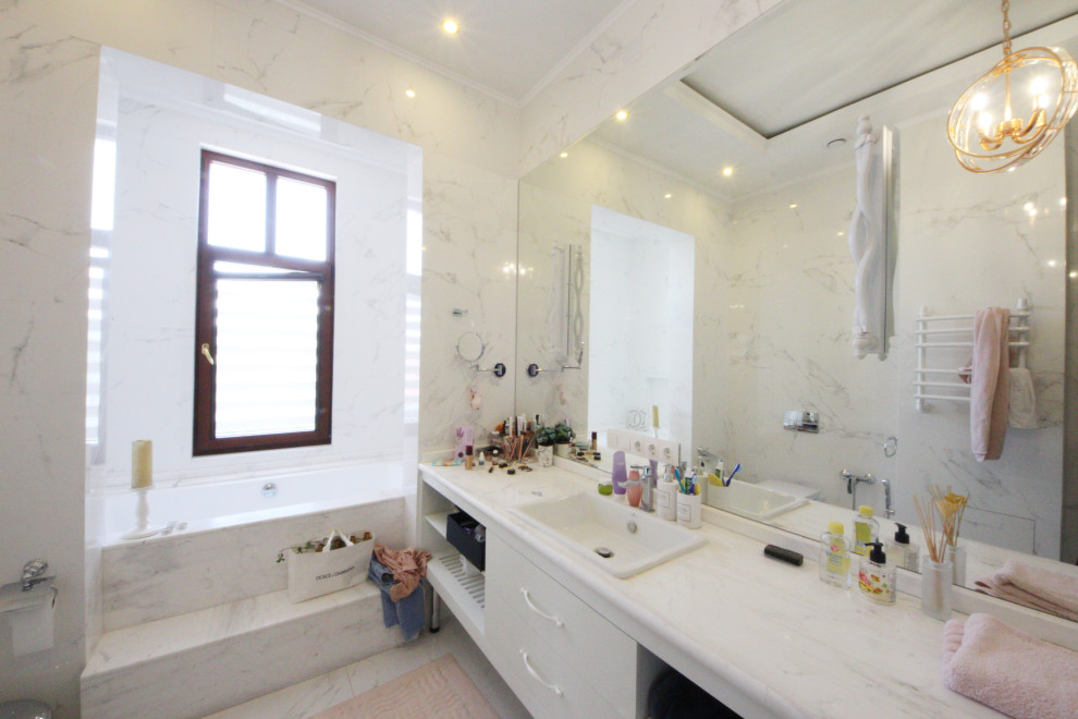 Eklektisk inredning av ett stort vit vitt badrum, med släta luckor, vita skåp, en vägghängd toalettstol, vit kakel, porslinskakel, vita väggar, klinkergolv i keramik, ett fristående handfat, marmorbänkskiva och vitt golv