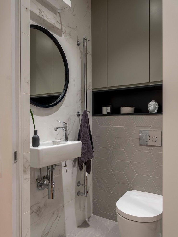 Modern inredning av ett toalett, med en toalettstol med separat cisternkåpa, vit kakel, porslinskakel, ett väggmonterat handfat, grått golv och grå väggar