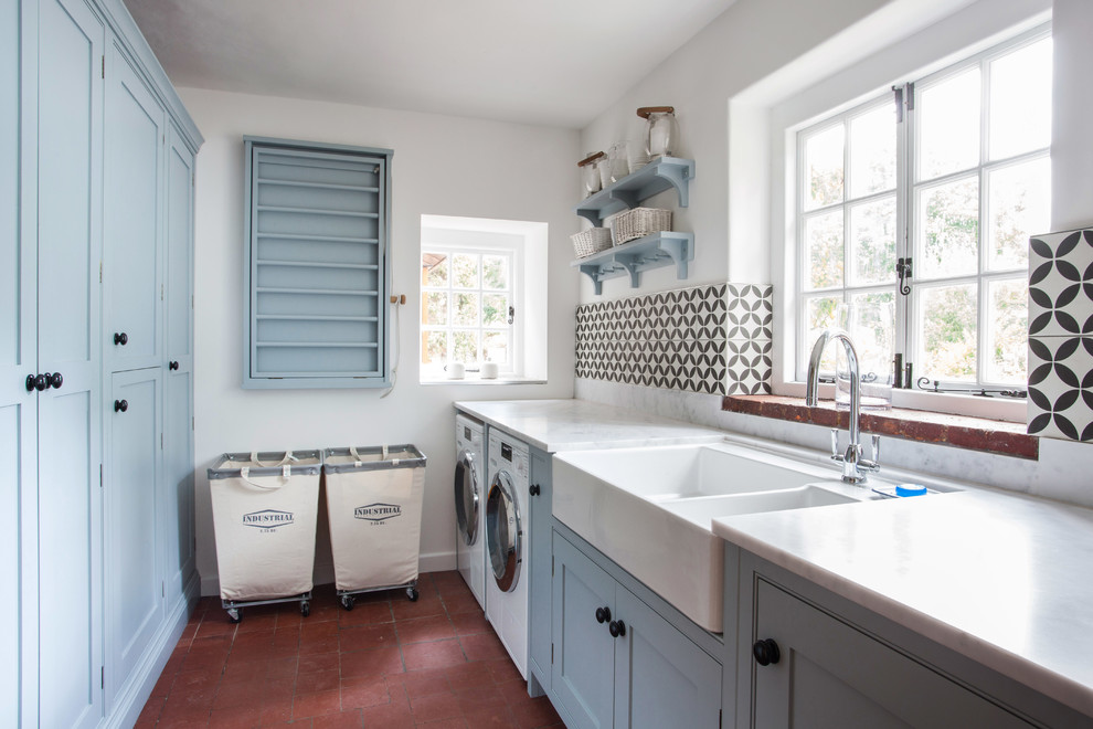 Idéer för att renovera en liten lantlig vita parallell vitt tvättstuga enbart för tvätt, med en rustik diskho, blå skåp, granitbänkskiva, vita väggar, klinkergolv i terrakotta, en tvättmaskin och torktumlare bredvid varandra, skåp i shakerstil och rött golv
