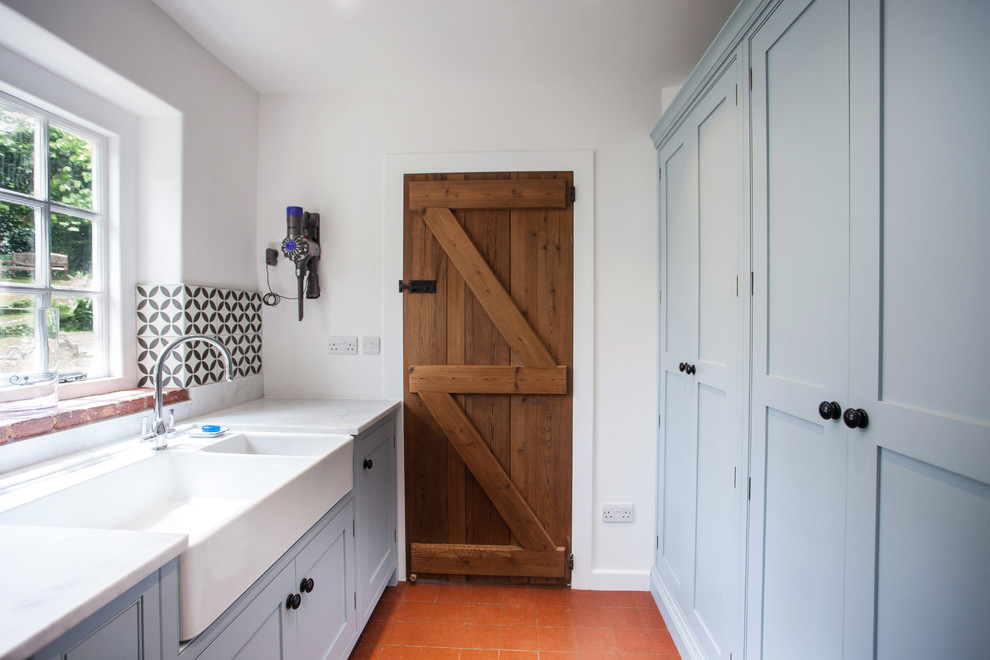 This is an example of a small farmhouse galley separated utility room in Kent with a belfast sink, recessed-panel cabinets, blue cabinets, granite worktops, white walls, terracotta flooring, a side by side washer and dryer, orange floors and white worktops.