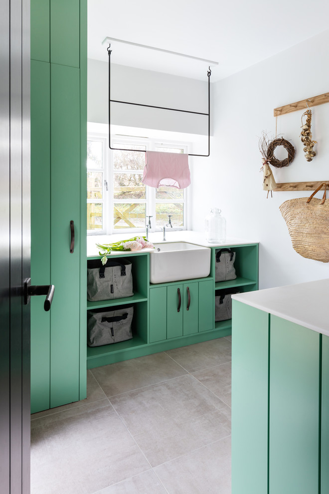 Example of a farmhouse gray floor laundry room design in Gloucestershire with flat-panel cabinets, green cabinets, white countertops, a farmhouse sink and white walls