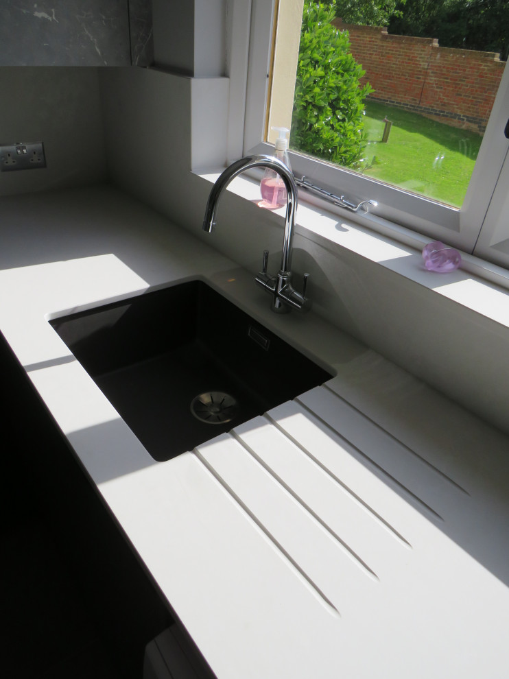 Inspiration for a medium sized modern l-shaped utility room in Essex with a submerged sink, flat-panel cabinets, green cabinets, quartz worktops, white splashback, engineered quartz splashback, grey walls, porcelain flooring, a side by side washer and dryer, grey floors and white worktops.