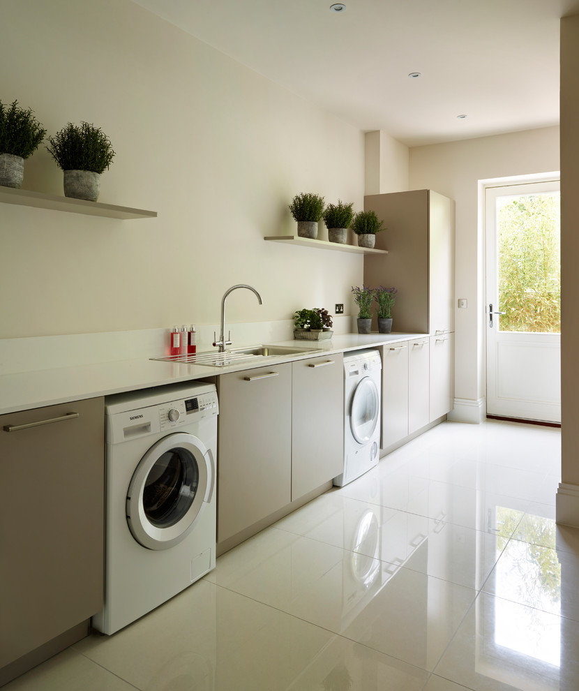 Ejemplo de lavadero lineal actual con armarios con paneles lisos, lavadora y secadora juntas, paredes beige, suelo blanco, puertas de armario grises y fregadero encastrado
