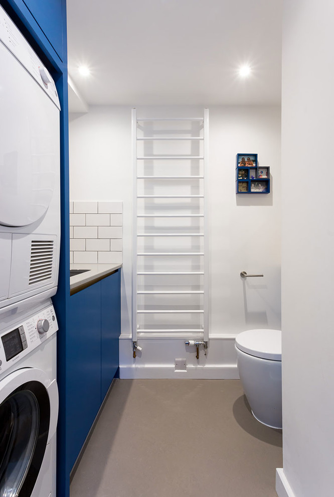 Cette image montre une petite buanderie parallèle design multi-usage avec un évier encastré, un placard à porte plane, des portes de placard bleues, un mur blanc, sol en béton ciré et des machines superposées.