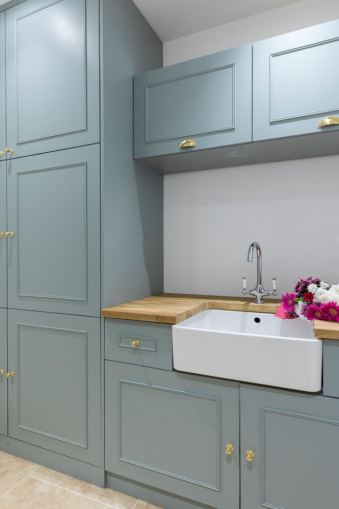 Medium sized rural single-wall utility room in London with a belfast sink, shaker cabinets, wood worktops, grey walls, porcelain flooring, a concealed washer and dryer, brown floors and blue cabinets.