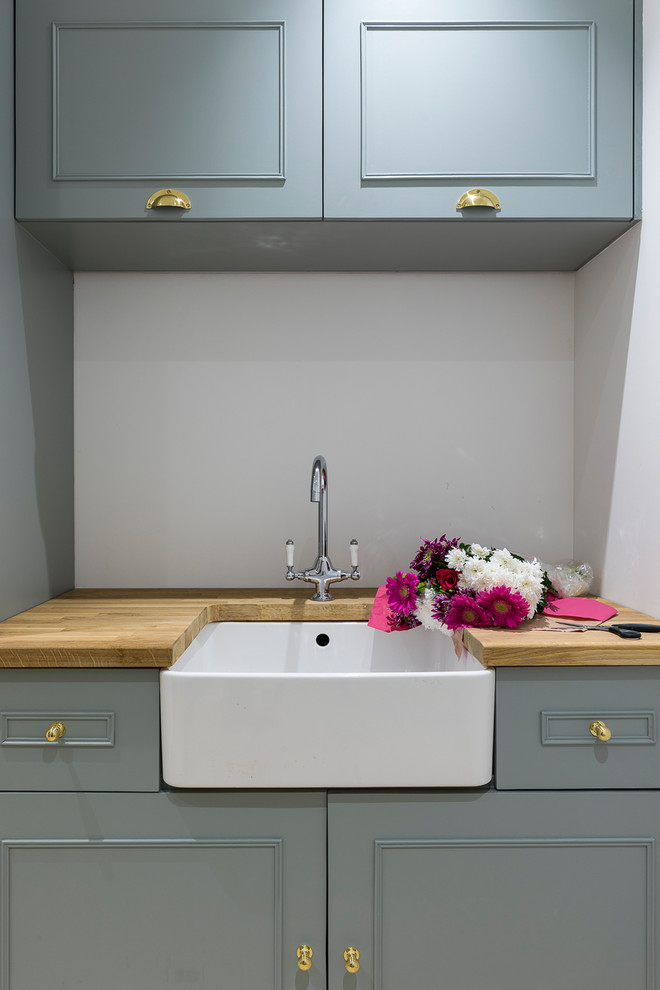 Medium sized farmhouse single-wall utility room in London with a belfast sink, shaker cabinets, blue cabinets, wood worktops, grey walls, porcelain flooring and a concealed washer and dryer.
