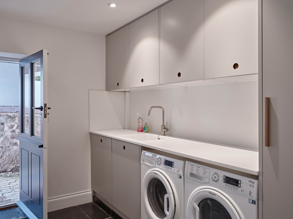 Foto di una lavanderia moderna con lavello da incasso, ante lisce, ante grigie, top in superficie solida, pareti bianche, pavimento in ardesia, lavatrice e asciugatrice affiancate, pavimento grigio e top bianco