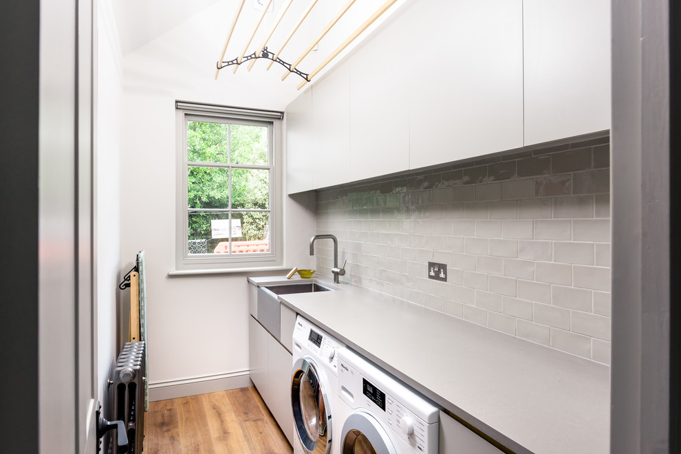 This is an example of a small contemporary single-wall separated utility room in Sussex with a belfast sink, flat-panel cabinets, grey cabinets, granite worktops, white walls, a side by side washer and dryer, grey worktops, medium hardwood flooring and brown floors.