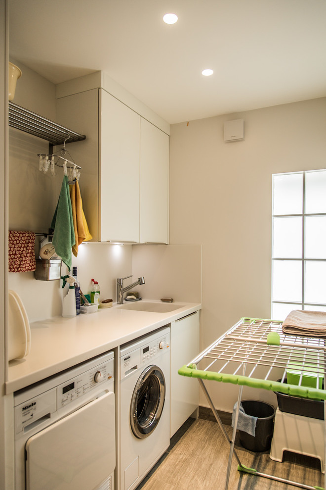 Zweizeilige Moderne Waschküche mit integriertem Waschbecken, flächenbündigen Schrankfronten, weißen Schränken, Quarzwerkstein-Arbeitsplatte, weißer Wandfarbe, Keramikboden und Waschmaschine und Trockner nebeneinander in London