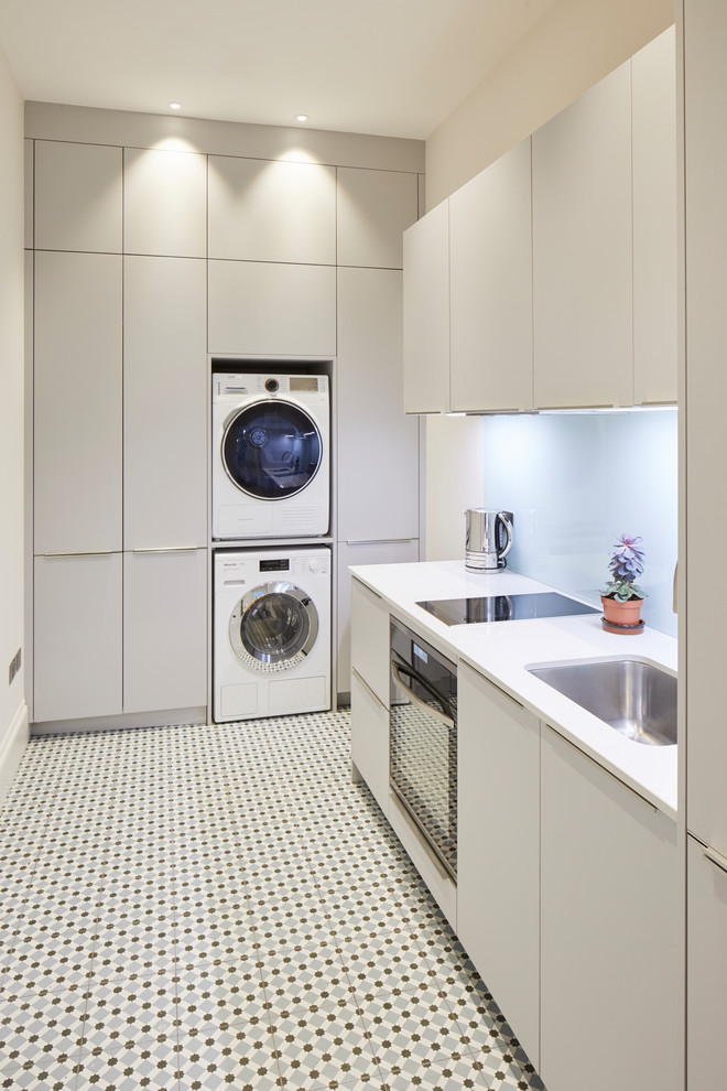 Multifunktionaler Moderner Hauswirtschaftsraum mit flächenbündigen Schrankfronten, grauen Schränken, weißer Wandfarbe, Waschmaschine und Trockner gestapelt und buntem Boden in London